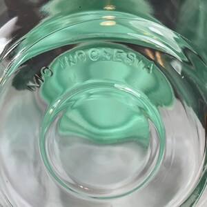 May include: Close-up of a clear, green-tinted glass object. The base features the embossed text "MG INDONESIA" in a circular pattern. The glass has a rounded, concave design.