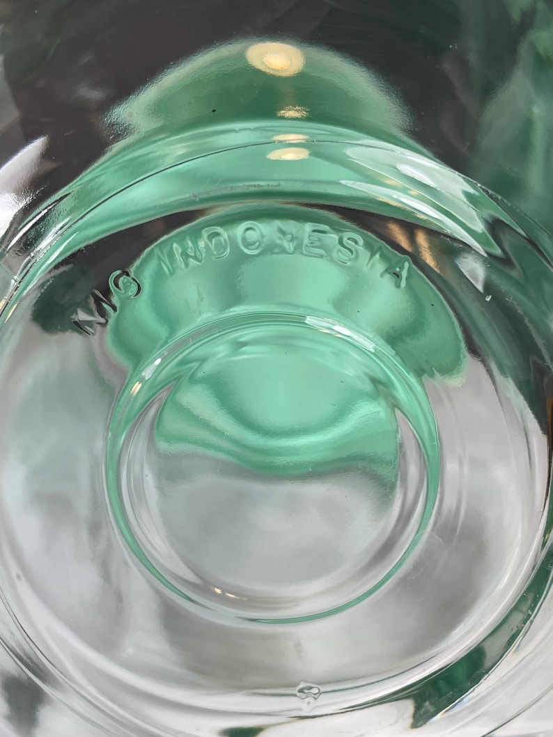 May include: Close-up of a clear, green-tinted glass object. The base features the embossed text "MG INDONESIA" in a circular pattern. The glass has a rounded, concave design.