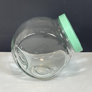 May include: A clear glass jar with a round, bubble-like shape and a light green lid. The jar has a wide opening and a slightly tilted design. The jar is empty and sits on a white surface with a dark background.