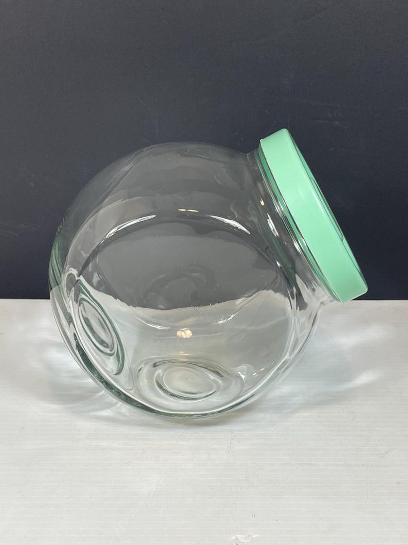 May include: A clear glass jar with a round, bubble-like shape and a light green lid. The jar has a wide opening and a slightly tilted design. The jar is empty and sits on a white surface with a dark background.