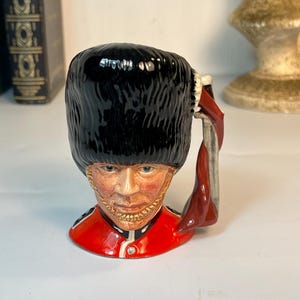 以下が含まれることがあります： 高い黒い熊皮の帽子と鮮やかな赤い制服のジャケットを着たイギリス兵の形をした陶器製のキャラクタージャグ。フィギュアは顎紐付きの精巧な顔立ちで、赤と白の制服のディテールがハンドルを形成しています。