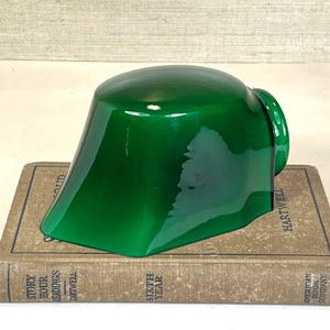 May include: A glossy, emerald green glass lampshade with a distinctive shape, placed on a vintage book. The lampshade has a rounded top and angled sides. The book has a tan cover with text. The lampshade is a decorative lighting element.