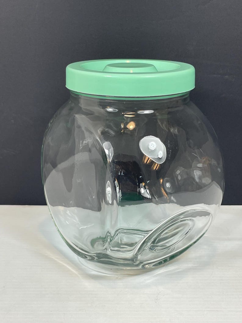May include: A clear glass jar with a rounded body and a light green lid. The jar has a wide opening and a smooth surface. The lid is a solid, opaque green color. The jar is empty and appears to be clean.