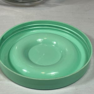 May include: A round, light green plastic lid with a raised, circular center. The lid has a series of concentric rings on the outer edge and a smooth, glossy finish. The lid appears to be for a container.