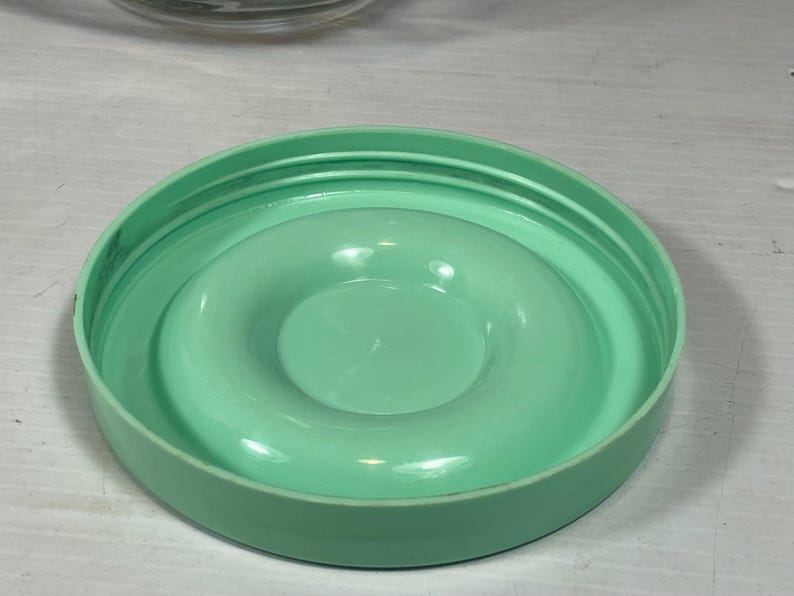 May include: A round, light green plastic lid with a raised, circular center. The lid has a series of concentric rings on the outer edge and a smooth, glossy finish. The lid appears to be for a container.