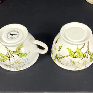 Op de afbeelding: Twee witte theekopjes van keramiek met een bloemenontwerp met witte bloemen en groene bladeren. De kopjes hebben een zwart vogellogo op de bodem met de tekst "John Austen and  Made in China".