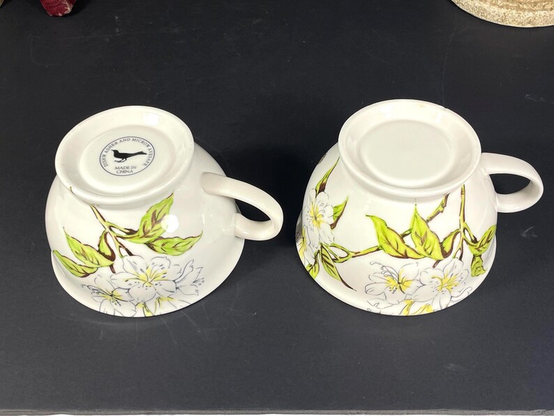 Op de afbeelding: Twee witte theekopjes van keramiek met een bloemenontwerp met witte bloemen en groene bladeren. De kopjes hebben een zwart vogellogo op de bodem met de tekst "John Austen and  Made in China".