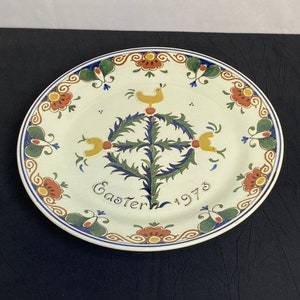 Könnte beinhalten: Ovale Keramikplatte mit cremefarbenem Hintergrund. Die Platte zeigt ein zentrales Design eines stilisierten Baumes mit gelben Vögeln und dem Text "Easter 1973". Der Rand hat Blumen- und Schmetterlingsmotive in Rot, Grün und Blau.