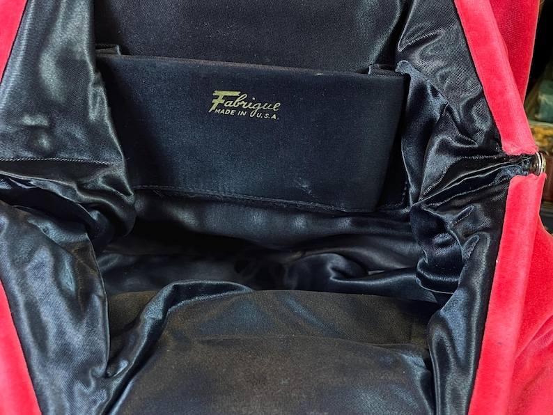 May include: A red velvet evening bag with a black satin lining. The bag has a zipper closure and a label that reads "Fabrique Made in U.S.A."