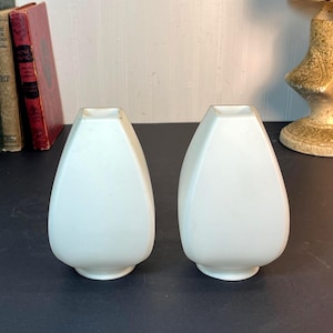 Puede incluir: Un par de jarrones de cerámica en color blanco mate con una silueta en forma de lágrima. Cada jarrón presenta una superficie lisa de color sólido, un cuello estrecho y una base ancha y redondeada, colocados como dos recipientes idénticos.