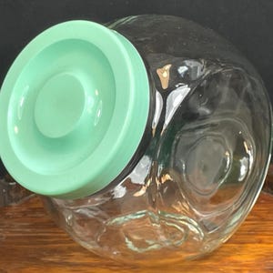 May include: A clear glass jar with a round, light green lid. The jar has a wide, rounded shape and appears to be empty. The lid is a solid, opaque color. The jar is sitting on a wooden surface.
