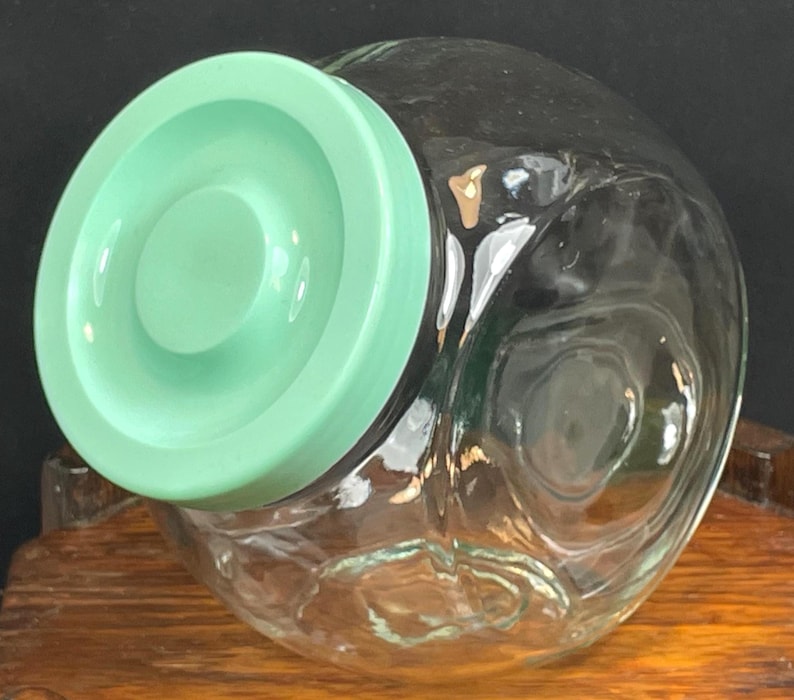 May include: A clear glass jar with a round, light green lid. The jar has a wide, rounded shape and appears to be empty. The lid is a solid, opaque color. The jar is sitting on a wooden surface.