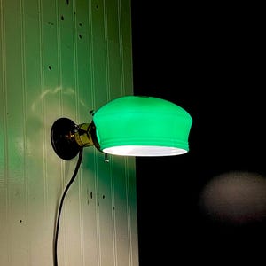 May include: A wall-mounted lamp with a bright green glass shade. The lamp has a brass-coloured arm and a black base, with a black cord. The shade emits a soft, green glow, casting light onto the white paneled wall.