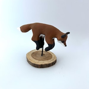 May include: A ceramic fox figurine in a jumping pose, painted brown with black accents, on a wooden base.