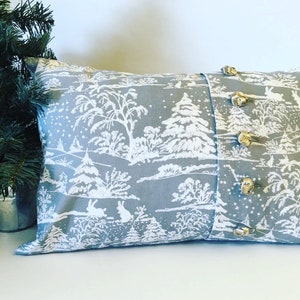 May include: A gray and white pillow with a winter scene design. The pillow has a white border and is decorated with gold jingle bells.