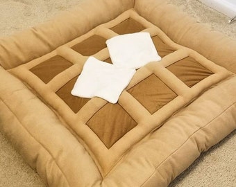 Puede incluir: Una cama para mascotas de color beige en forma de gofre con dos almohadas blancas. La cama está hecha de tela suave y felpa y tiene un borde elevado para mayor comodidad.
