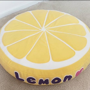 May include: A round, yellow cushion shaped like a lemon slice. The cushion has white accents and the word "Lemon" in purple letters. A pink heart is next to the word. The cushion is on a light-colored carpet.