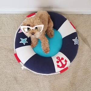 May include: A small brown dog wearing pink sunglasses sits in a blue and white life preserver shaped dog bed. The bed has a red anchor and white stars on it.