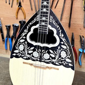 Peut inclure: Un instrument à cordes traditionnel, probablement un bouzouki, avec un corps crème et une face noire ornée de motifs floraux blancs. L'instrument est présenté sur un fond d'outils divers, dont des tournevis et des pinces.