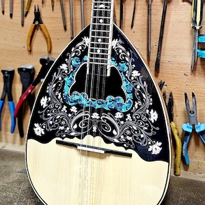 Könnte beinhalten: Ein traditionelles Saiteninstrument mit birnenförmigem Korpus, schwarz-weißem Blumendesign und hellem Resonanzboden. Das Instrument wird in einer Werkstattumgebung ausgestellt, umgeben von Werkzeugen.