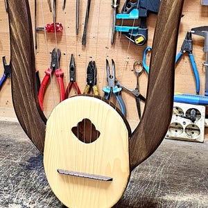 Puede incluir: Una lira de madera hecha a mano con una caja de resonancia de color claro y brazos curvos de color marrón oscuro. El instrumento se exhibe en un taller, con varias herramientas colgando en la pared detrás.