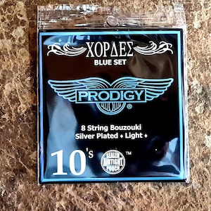 May include: A pack of Prodigy 8-string bouzouki strings in a blue set. The pack is black with a blue and white logo and the text "Χορδές Blue Set" and "Prodigy". The pack also says "8 String Bouzouki Silver Plated + Light + 10's".
