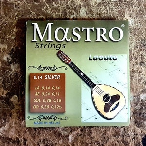 Pode incluir: Um pacote de cordas prateadas para um laouto, um tipo de alaúde. O pacote é etiquetado como "MASTRO Strings" e inclui a calibre de cada corda. O pacote também apresenta uma imagem de um laouto.