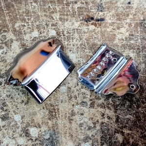 May include: Two chrome-plated metal guitar bridge covers. The covers are on a worn wooden surface.