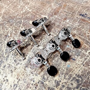 May include: Four silver guitar tuning pegs with black knobs. The pegs have a decorative design.