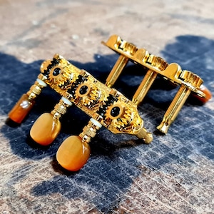 May include: A gold-coloured guitar tuning machine head with intricate detailing. The headstock features decorative black accents and amber-coloured tuning pegs. The hardware is set against a textured, wooden surface.