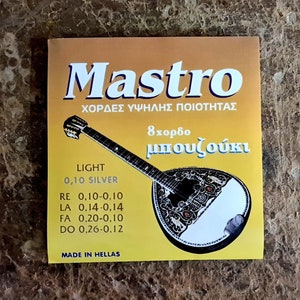 Puede incluir: Un paquete de cuerdas de bouzouki Mastro. El paquete dorado y blanco muestra el nombre de la marca y las especificaciones de las cuerdas. El texto incluye "LIGHT 0,10 SILVER" y medidas para las cuerdas RE, LA, FA y DO. Hecho en Grecia.