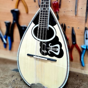 May include: A wooden mandolin with a black and white design on the body. The mandolin has a light wood finish and a black fretboard. The mandolin is sitting on a wooden surface.
