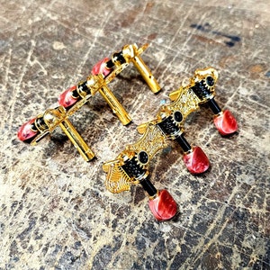 May include: Close-up of several gold-colored guitar tuning machines. Each machine has a decorative design, black accents, and red tuning pegs. The machines are arranged on a textured, neutral-toned surface.