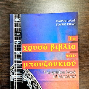 Könnte beinhalten: Ein Buchcover mit rotem, orangem und blauem Hintergrund. Der Titel lautet "The Golden Book of Bouzouki" in Englisch und Griechisch. Das Cover zeigt ein schwarz-weißes Bouzouki mit aufwendigen floralen Designs.