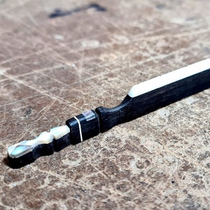 Puede incluir: Un puente de guitarra negro y blanco con una incrustación blanca. El puente está hecho de madera y tiene un acabado liso.