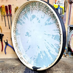 Può includere: Un grande tamburo rotondo fatto a mano con una superficie a motivi blu chiaro e bianca. Il tamburo ha un telaio in legno con un bordo nero ed è decorato con piccoli anelli di metallo. Vari strumenti sono appesi alla parete di legno dietro il tamburo.