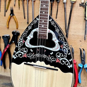 Peut inclure: Un bouzouki, instrument à cordes traditionnel, avec un corps en bois clair et un motif décoratif noir et blanc. Divers outils, dont des tournevis, des pinces et des pinceaux, sont disposés sur le mur en bois derrière l'instrument.
