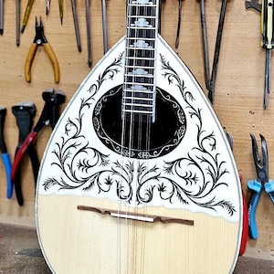 May include: A pear-shaped, light-colored stringed instrument with a decorative black and white design. The instrument is surrounded by various tools, including screwdrivers, pliers, and brushes, all hanging on a wooden wall.