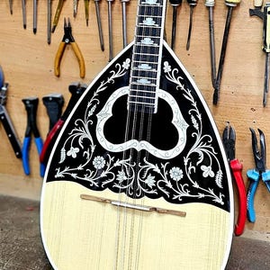 Peut inclure: Un bouzouki grec traditionnel avec un corps crème et un motif décoratif noir et blanc. L'instrument est exposé dans un atelier, entouré de divers outils.