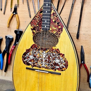Op de afbeelding: Een houten bouzouki met een gele body, een zwart toetsbord en ingewikkelde bloemmotieven. Het instrument hangt aan een muur met verschillende gereedschappen en borstels.
