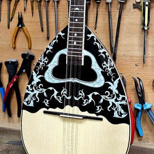 May include: A traditional stringed instrument with a pear-shaped body, black and white design, and intricate floral patterns. The instrument is displayed against a wooden wall with various tools, including screwdrivers, pliers, and brushes.