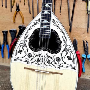 Peut inclure: Un instrument à cordes traditionnel, probablement un bouzouki, avec un corps en bois clair et un motif décoratif noir et blanc. L'instrument est entouré de divers outils, dont des tournevis, des pinces et des pinceaux, suggérant un atelier.