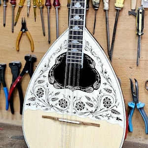 May include: A traditional stringed instrument, likely a bouzouki, with a cream-colored body and a decorative floral design. The instrument is displayed against a wooden wall with various tools, including screwdrivers, pliers, and brushes.