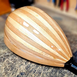 Pode incluir: Um instrumento musical de madeira com um corpo em forma de p&ecirc;ra e um pesco&ccedil;o preto. O corpo &eacute; feito de tiras de madeira clara e escura, criando um padr&atilde;o listrado.