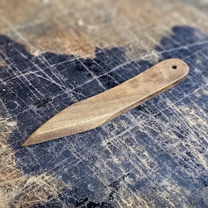 Può includere: Un coltello da lancio in legno con una punta arrotondata e un foro vicino al manico. Il coltello è appoggiato su una superficie di legno usurata.