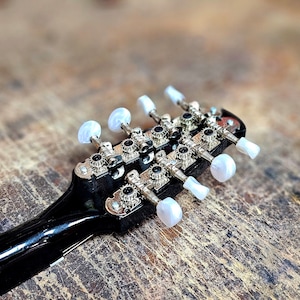 Pode incluir: Close-up da cabe&ccedil;a de um bandolim preto com pinos de afina&ccedil;&atilde;o prateados e bot&otilde;es de pl&aacute;stico branco.