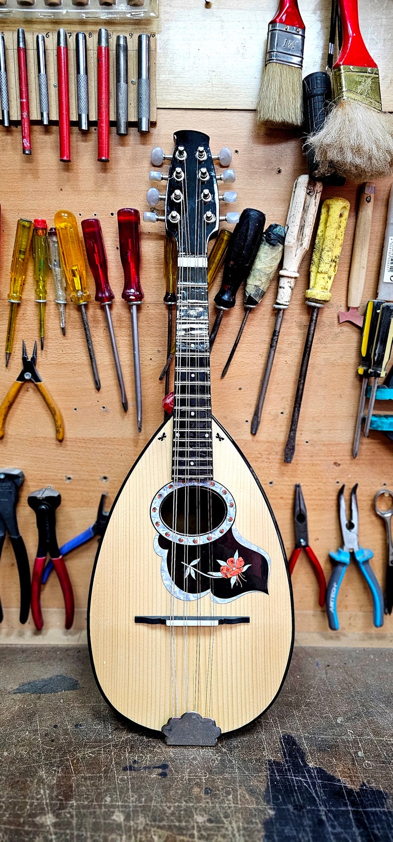 Pode incluir: Um bandolim de madeira com um desenho floral preto e branco na caixa de resson&acirc;ncia. O bandolim est&aacute; rodeado de v&aacute;rias ferramentas, incluindo chaves de fenda, alicates e pinc&eacute;is.