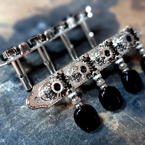 May include: Close-up of a set of silver-colored guitar tuning pegs with black knobs. The pegs are mounted on a silver plate with decorative details.