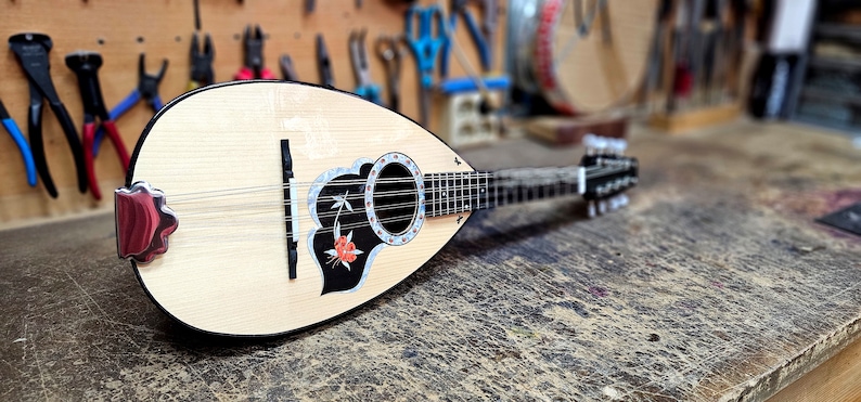 Pode incluir: Um ala&uacute;de de madeira com um desenho floral preto e branco na caixa de resson&acirc;ncia. O ala&uacute;de tem um bra&ccedil;o preto e pinos de afina&ccedil;&atilde;o prateados.