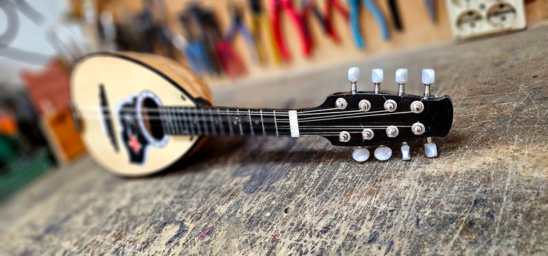 Pode incluir: Um bandolim de madeira com um corpo marrom claro e um bra&ccedil;o preto. O bandolim tem pinos de afina&ccedil;&atilde;o brancos e est&aacute; apoiado em uma superf&iacute;cie de madeira.